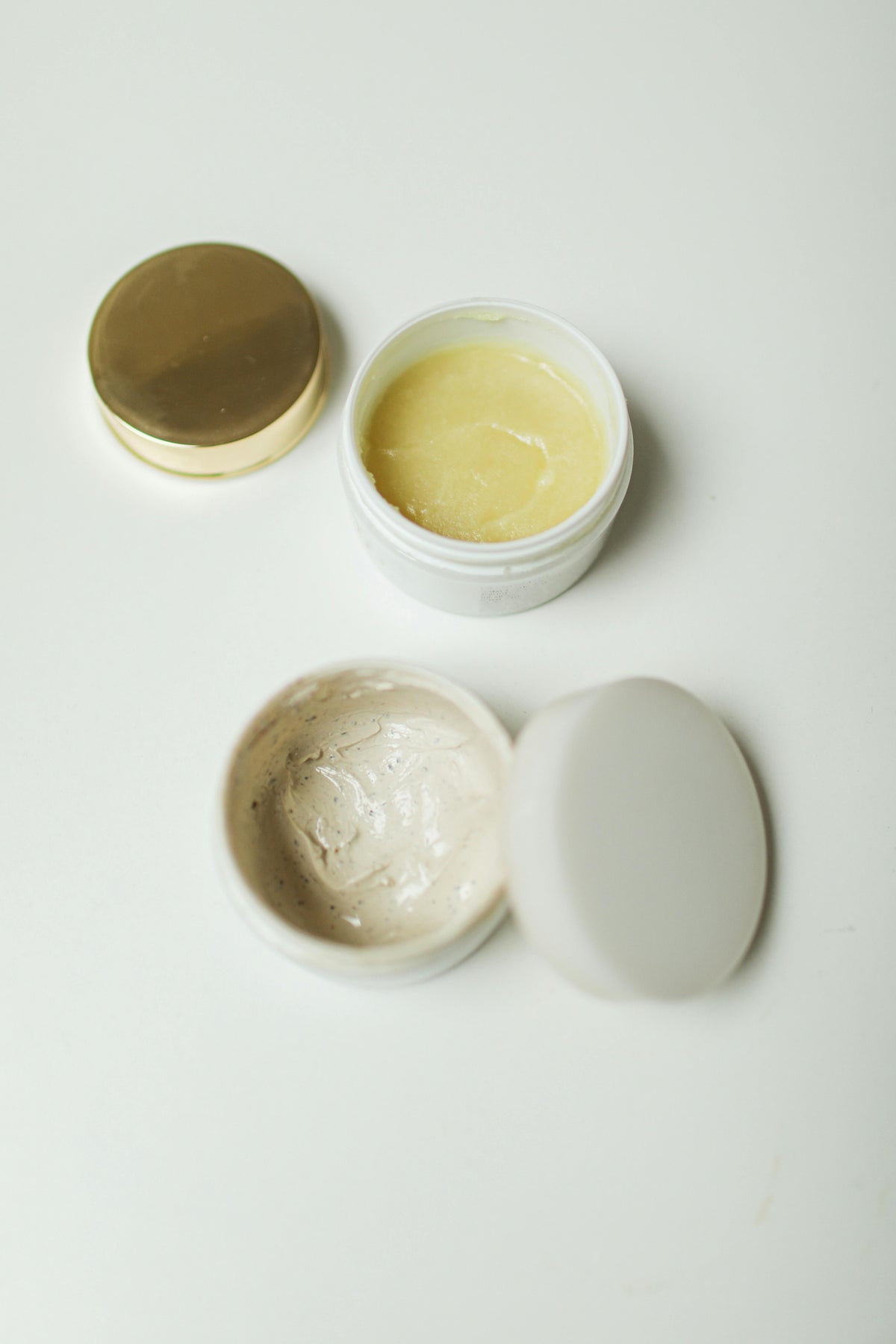 Three natural skincare butters in open glass jars on a neutral surface, illustrating a comparison of tallow, coconut oil, and lanolin